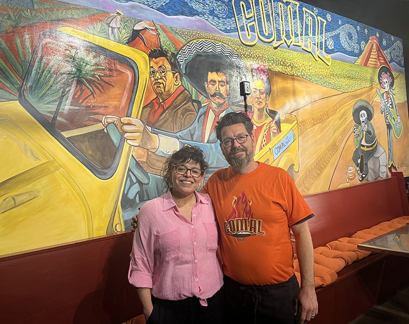 Two people stand in front of a mural. The mural features artists Frida Kahlo and Diego Rivera with Mexican Revolutionary Zapata.