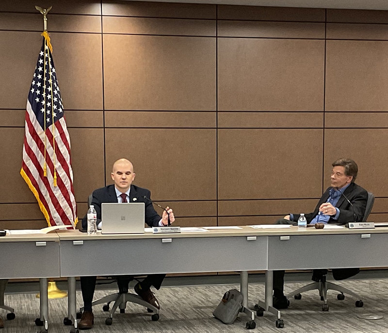 Two men, seated at a meeting table at the front of a room.