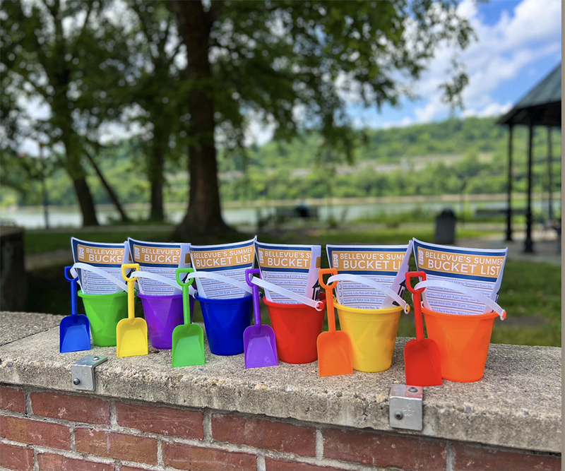 A row of colorful plastic buckets or pails with plastic shovels lined up along a low brick wall. The buckets have papers stuffed inside them.