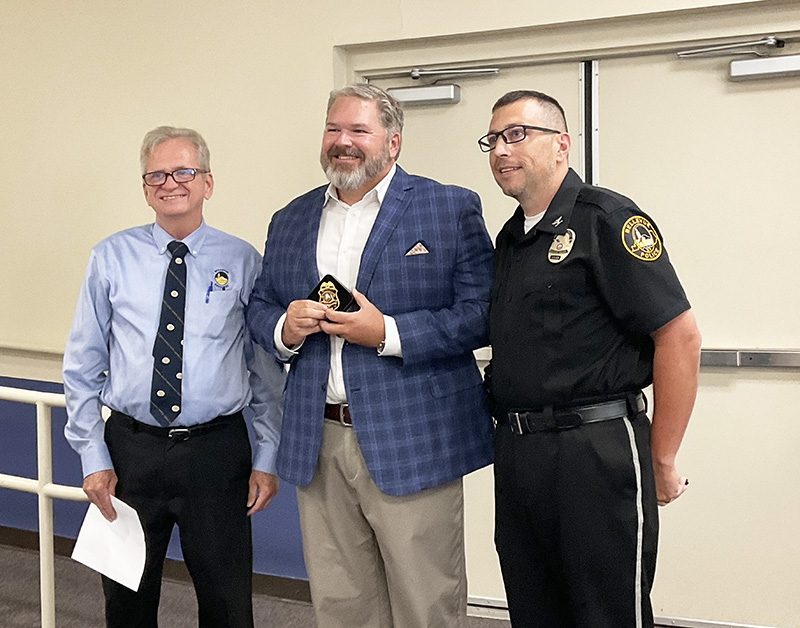 Three men stand. One is holding an award. Another is dressed in a police uniform.