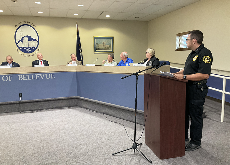 A police chief stands at a podium in front of a dais with six people seated. A city seal and flag is behind the dais.