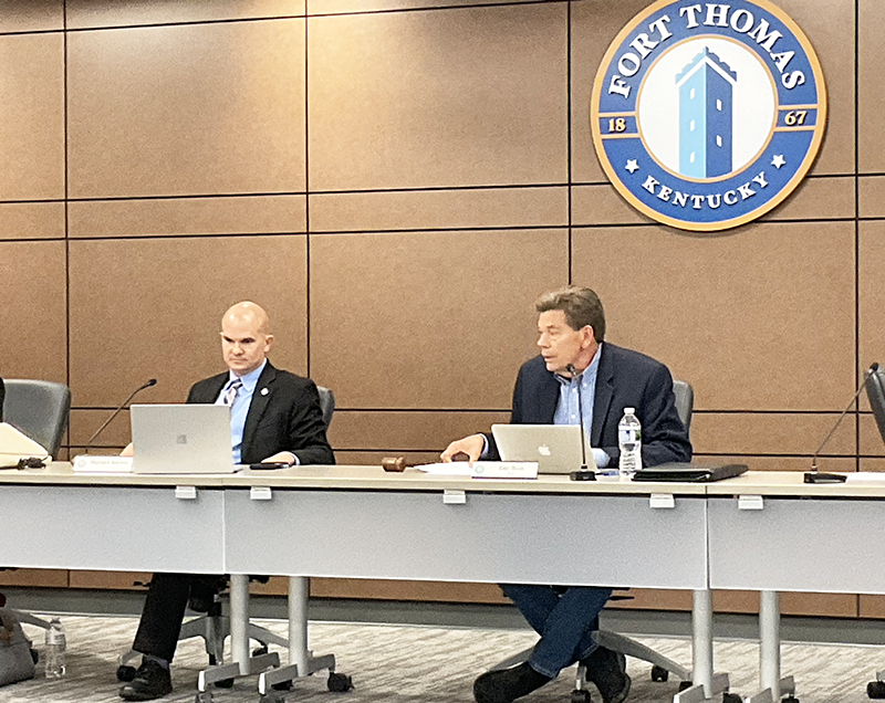 Two men seated at a dais with a seal of the city on the wall behind them.