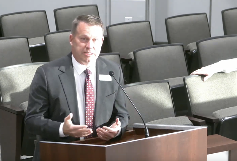 Man in a suit at podium is talking. There are empty seats around him.
