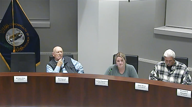 Two men and a woman sit behind a dais. There is a Kentucky state flag behind them.