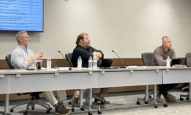 Three men sit behind a dais. On the wall behind them is a computer screen.