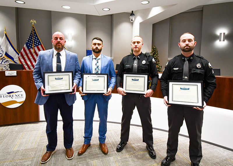 Four men stand in front of a dais holding certificates. Two are dressed in police uniforms.