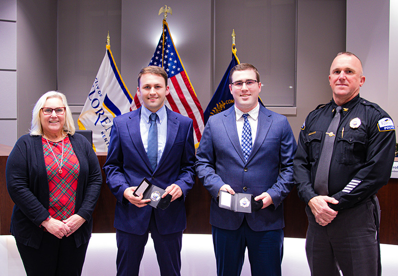 Four people standing in front of an American flag. Two men in the middle are holding police badges.