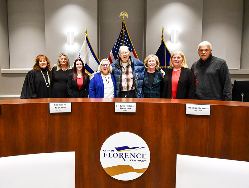 A group of eight people stand behind a dais bearing the city of Florence, Kentucky, seal. Behind them is the American flag, Commonwealth of Kentucky flag and city of Florence, Kentuck, flag.