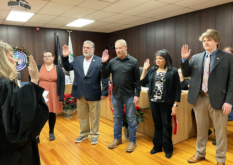 A group of 3 men and 2 women raise their hands as a judge in a black robe administers an oath of office.