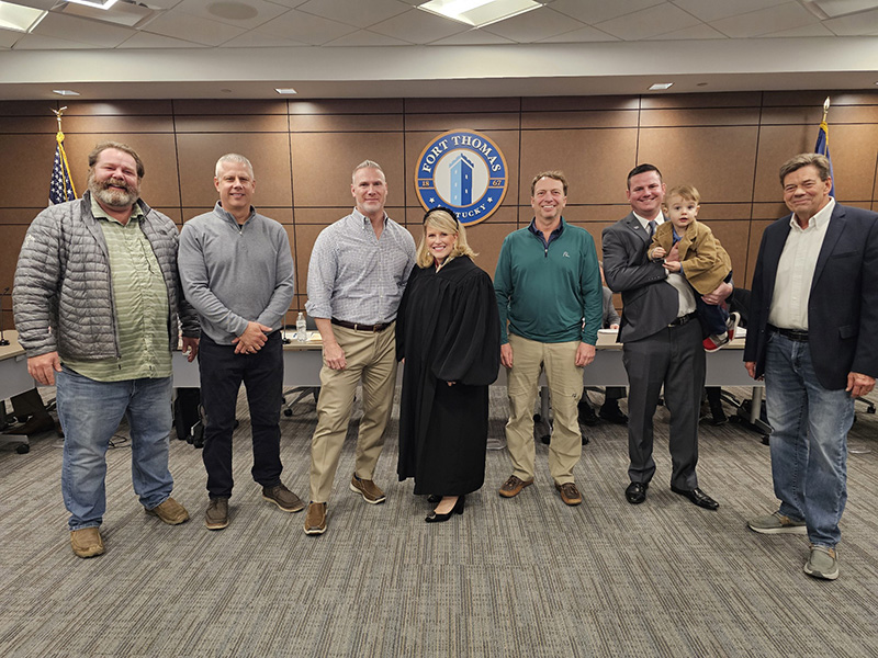 A group of six people stand with a judge dressed in a black robe. Behind them is a dais with the city of Fort Thomas seal on wall behind.