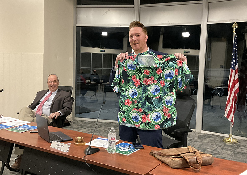 A man standing behind a dais holds up a Hawaiian shirt. A man seated next to him is laughing.
