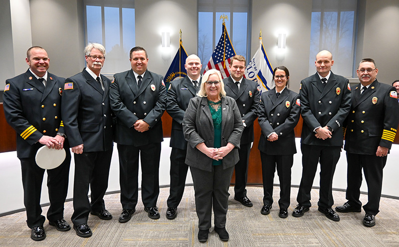 A group of 8 people dressed in formal firefighter uniforms stand behind a woman in business attire.