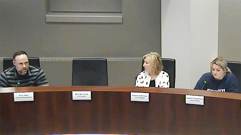 Three people sit at a dais. One man, who is speaking, is at the left next to an empty chair and two women sit to the right of the chair are listening.