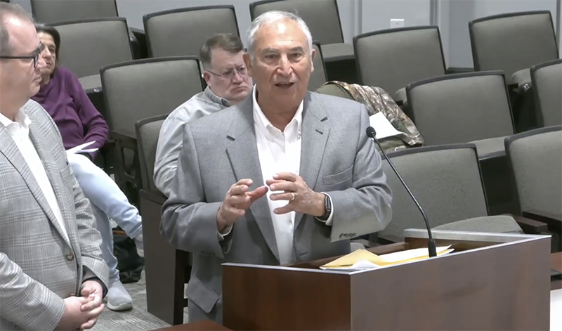 Two men stand at a podium. One is speaking and gestering with his hands.