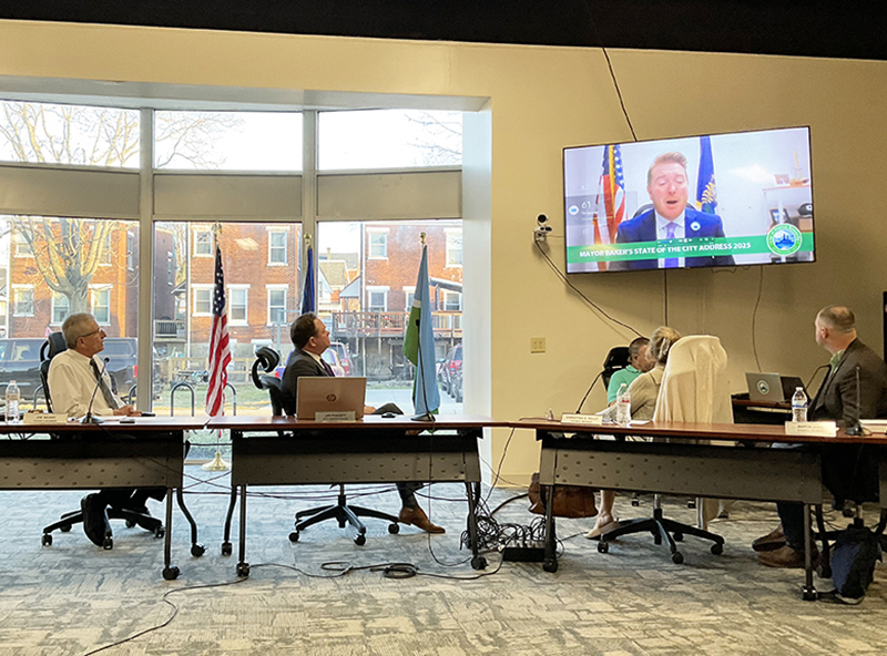 Four people sitting behind a dais have turned their chairs to watch a video overhead. A man is talking on the video screen.