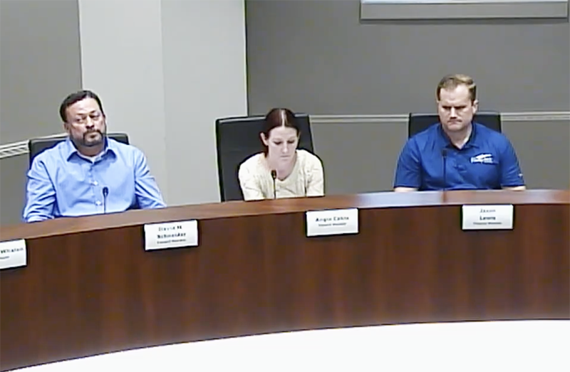 Three people sit at a dais. The woman in the middle is speaking; two men are listening.