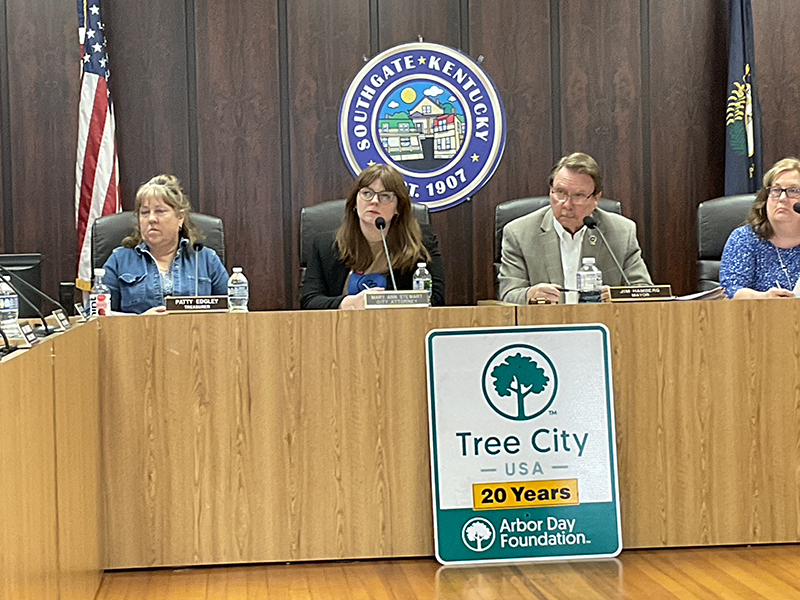 Four people sit at a dais. Behind them is an American flag, a Kentucky flag and the seal of the city of Southgate.