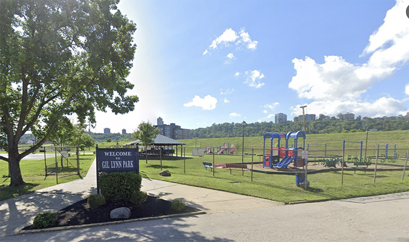 A park and playground. There is a sign "Gil Lynn Park" at the edge of the park, under a tree.