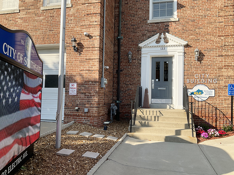 Front of a building with a sign "City Building" next to the front door and an electronic sign on the side. The electronic sign shows a waving American flag image.