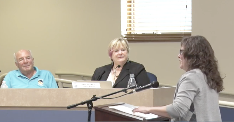 A woman stands at a podium addressing two people seated behind a dais
