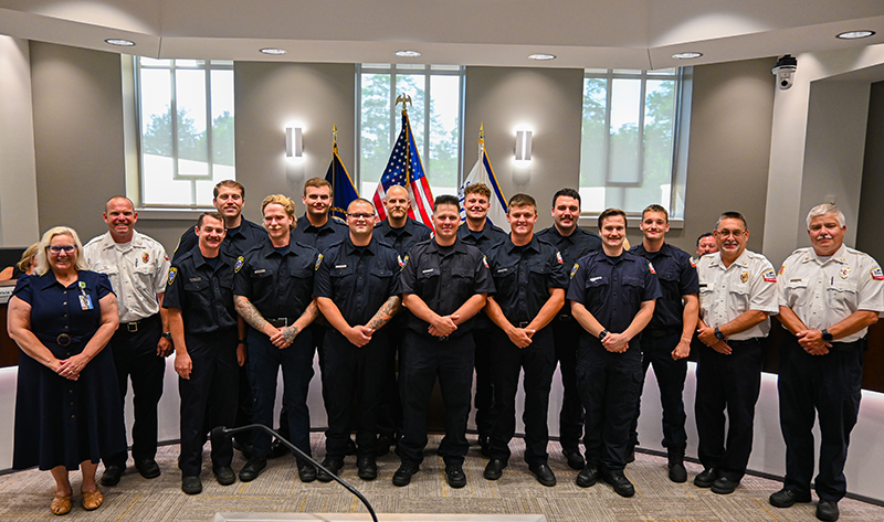 A group of 16 people pose in two rows for a photo. All but one are wearing fire service uniforms.