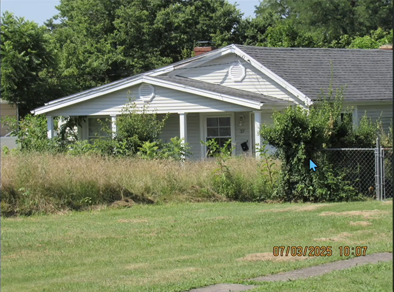House with tall grass and weeds in the front yard and tall weeds spilling over from the back yard.