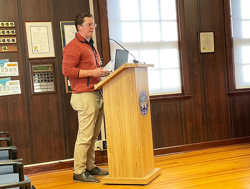 Man stands at a podium. City plaques are on the wall behind him.