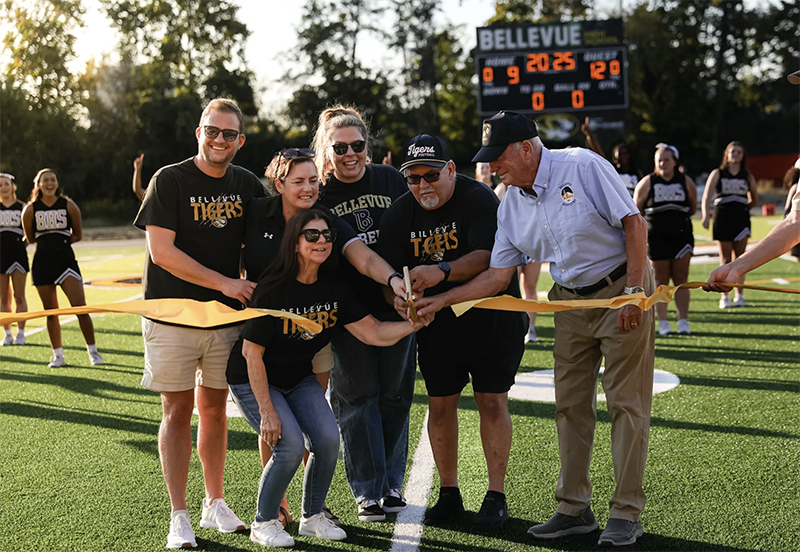 Group of 6 people cutting a ribbon on a football playing field. Five of the people are wearing Bellevue Tigers shirts and the sixth is wearing a coach's shirt and cap.