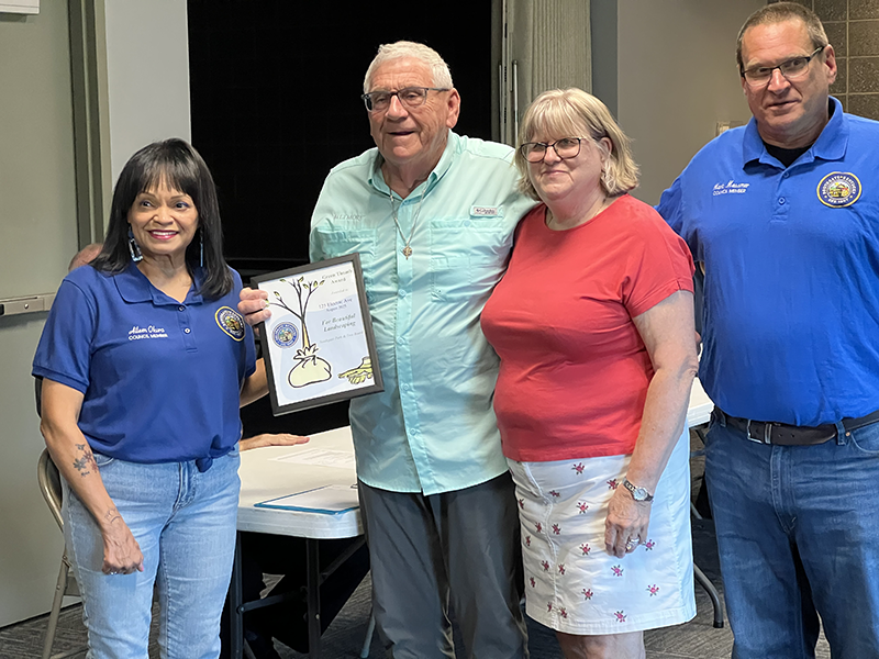 Four people pose for a photo. In the center, a man and woman pose with a certificate. The people on each end are wearing identical city official shirts.