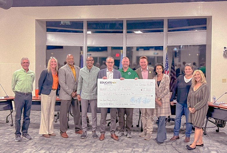 A group of 10 people, men and women, stand in front of a dais. The man in the center is holding a large check.