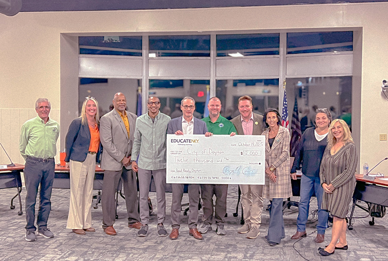 A group of 10 people, men and women, stand in front of a dais. The man in the center is holding a large check.
